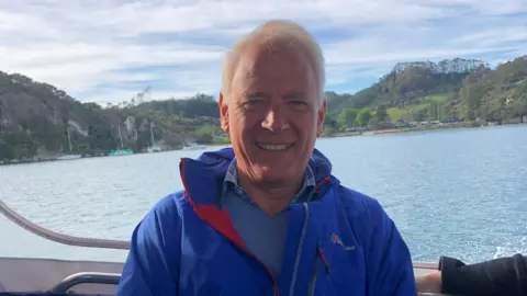 A man in a blue jacket is smiling while sat on a boat. Water and green hills can be seen in the background.