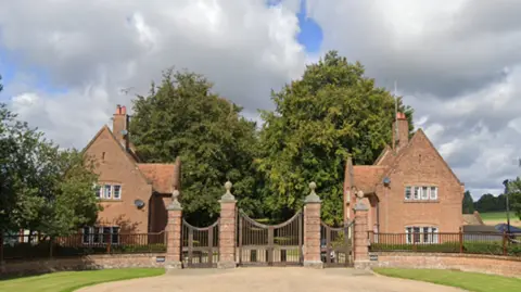 The gates at the entrance to the Chequer's estate. Three large imposing gates with red-brick houses on either side of the entrance. Before the gate is gravel, with banks of grass on either side. 