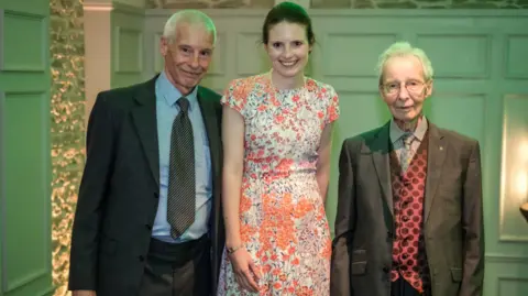 Rebecca Hillier Rebecca flanked by her father and grandfather in a large elegant room. She is wearing a floral peach summer dress and her relatives are in smart suits. Her grandfather is also wearing a red polka dot waistcoat. They are smiling to camera.