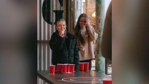 Factory Floor A woman with long blonde hair wearing a black coat aims a white ball in a game of beer pong inside a new venue inside an old cotton mill. Another woman stands to the side of her, watching her take aim. She has long dark brown hair and wears a beige top. They are both laughing.