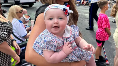 A baby is wearing a hairband that says my first 12th of July. She is wearing a dress with a blue floral pattern. A woman with brown hair is holding her. A crowd is in the background.