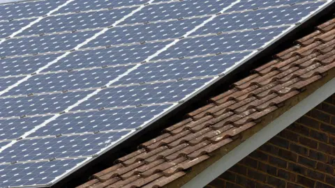 Close-up of a sloped house roof fitted with rows of blue solar panels mounted above brown tiled roofing.
