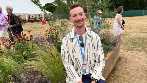 Clare Worden/BBC A man is sitting on the edge of a wooden garden planter. Behind him is a pine tree and in front of that is a range of grasses and flowers. The man is wearing a white shirt with blue stripes on it. He has brown hair. 
