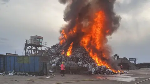 A firefighter dealing with a blaze at a recycling centre. A large plume of smoke is filling the sky directly above a pile of rubbish.