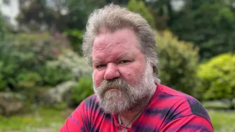 A man with wild grey hard and a great big bushy beard with a red and blue shirt.