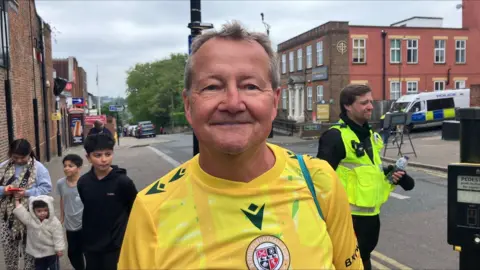Simon Elwell smiling looking at the camera, on a pavement, with a family of one mother and three boys and a police officer in the background