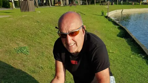 Glenn Orrah A man wearing sunglasses looks at the camera. He is crouching next to a fishing lake.