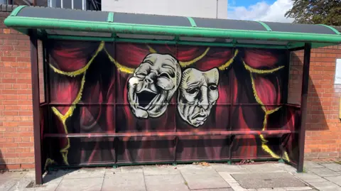 Nue/Wingy A bus stop designed like a theatre with red curtains and yellow trim. With a smiling mask and sad mask face in the centre. 