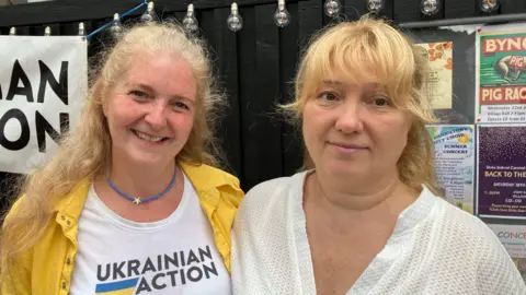 Two people stand side by side outdoors in front of a black wooden fence decorated with string lights and community event posters. One person wears a yellow jacket over a white T‑shirt that reads “Ukrainian Action” and a blue necklace with a star-shaped pendant. The other wears a white textured top. Both face the camera.
