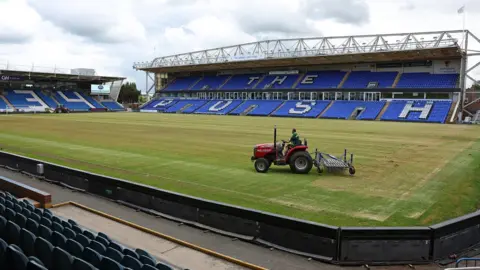 A view of Peterborough United's stadium, with a man riding a small red tractor across the pitch. The tractor has a rake attachment at the back. The stand opposite has blue seating, with The Posh spelt out in white seating.