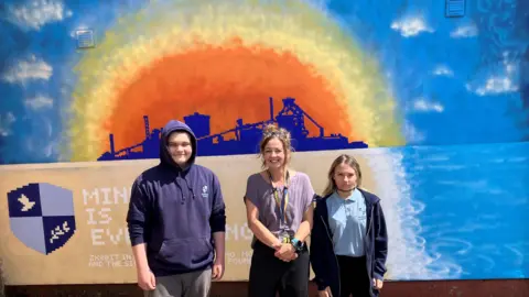 Teacher Carla Hindle standing in front of the steelworks mural, with a boy on her right side and a girl to her left. The artwork shows the former factory in silhouette within an orange sun. It is surrounded by blue sky.