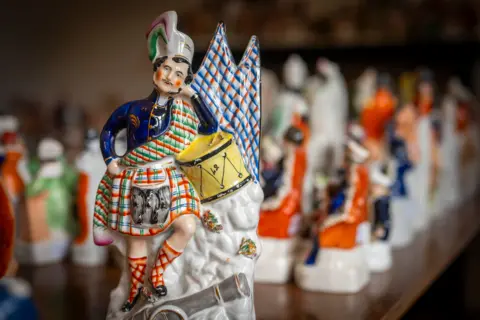 Phil Wilkinson A ceramic Highland soldier with a drum stands on a cannon in front of  a range of other pottery items