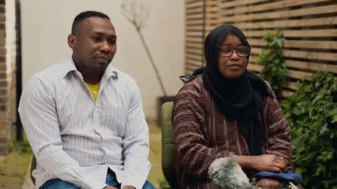 Mr Mohammed is sat on the left, wearing a striped shirt with a yellow top underneath and Mrs Mohammed, who is sat next to her husband, is wearing glasses, a hijab and a long red top.