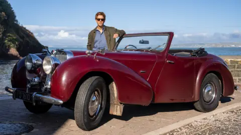 UKTV Damien Molony standing by the door of a red Triumph Roadster