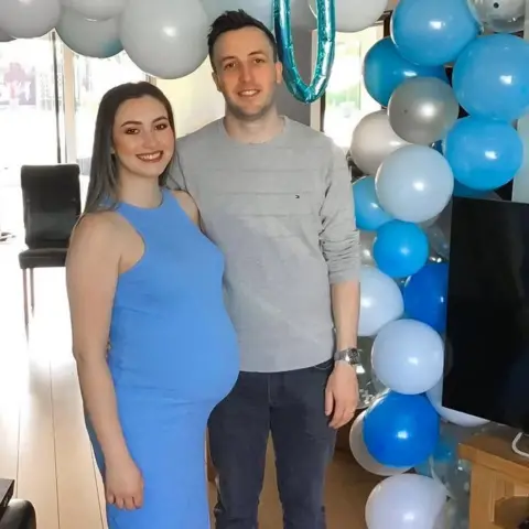Kianty and Conor Simpson Kianty in a blue dress with a visible baby bump is smiling next to Conor. There's a balloon arch with white and pale and dark blue balloons behind them.