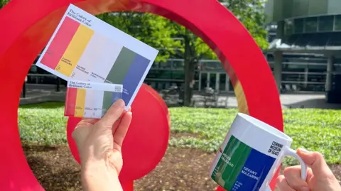 Corning Museum of Glass Two postcards with colour strips of red, yellow, ivory, green and blue being held in one hand and a white mug with green and blue stripes in the other.