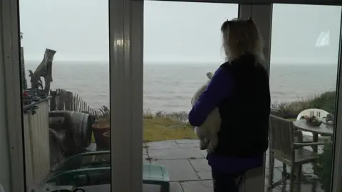 Martin Giles/BBC Hilary holds her dog in her arms as she looks out of her French windows towards the sea. It is a grey and foggy day. 