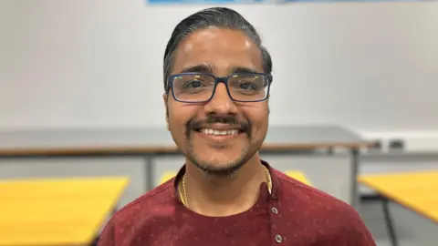 JOANNA TAYLOR/BBC Jeetendra smiling at the camera. He is wearing glasses, a gold chain and a red shirt. An out of focus classroom is in the background. 