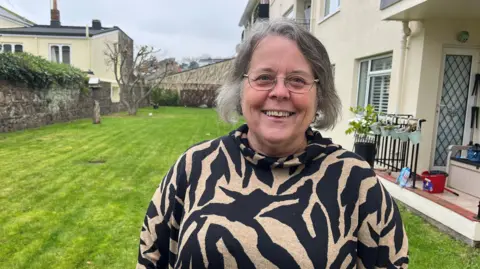 BBC Jeannine Carey standing in a garden with a white building in the background. She is looking into the camera, while wearing an animal print top. She has short grey hair and glasses. 