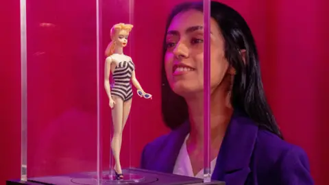 PA Media Fatima Zahra Zeyani looks at the very first Barbie doll from 1959 on display during a preview of Barbie: The Exhibition at Kelvingrove Art Gallery and Museum in Glasgow. The figure is in a glass case.