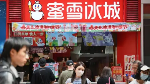 A shop of Mixue, a Chinese bubble tea brand in Hong Kong in March 2026. In the foreground a crowd passes by the shop.