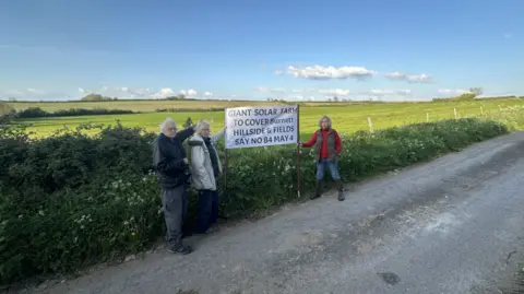 Two women holding a sign saying 'giant solar farm to cover Burnett hillside and fields, say no b4 May 4'. An elderly man is stood close to the sign, which is being held up by an empty field. There are other fields in the background and blue sky.