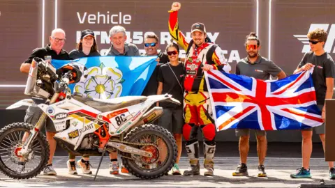 Robbie Wallace is in the centre of the image surrounded by teammates and family and his motorbike is to the left of the image. The group is holding a Union flag and a Yorkshire flag