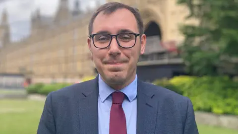 Liberal Democrat MP for Harrogate and Knaresborough standing outside the Houses of Parliament, Tom is wearing black framed glasses, a light blue shirt with a red tie and a navy blazer.