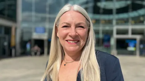 JO THEWLIS/BBC Kay Mason Billig has long, blonde hair and is wearing a blue jacket.
She is smiling at the camera with a glass building, which is out of focus, behind her.