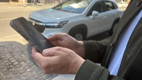 A man stands in a street holding up an Iphone. A car is parked behind him. 