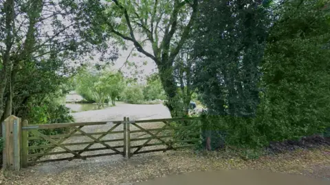 A pond and nature area seen from a road. A wooden double gate bars the entrance. Trees and bushes can be seen lining the site and by the pond. Leaves are strewn on the ground.