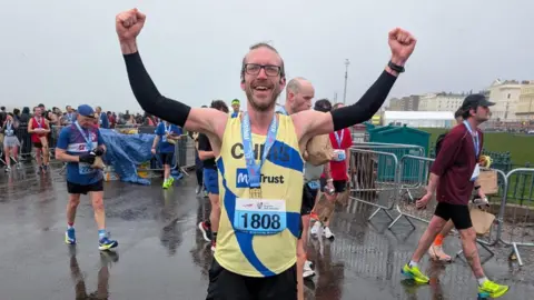 Chris White stands on a wet road at the finish area of a race, holding both arms in the air. He is wearing a yellow and blue race vest, black shorts, arm sleeves, and a medal around the neck. Other participants and spectators move around in the background near metal barriers.