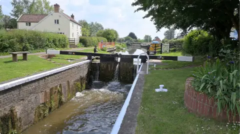 A canal is seen stopped by a lock. There is grass next to the canal and some houses.