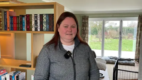 A woman wearing a grey zipped hoodie over a white T-shirt. She has long brown hair and is standing in a room in her house with a patio and grass in the background.