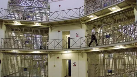 The inside of a prison where a guard walks along on the second floor of a rotunda surrounded by barred cells. 