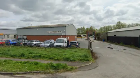 A small industrial estate with a low, grey warehouse labelled “Lugg Valley Primrose Travel.” Several parked cars line the fenced front area. To the right, a narrow road leads past another warehouse, with a couple of buses visible in the background.