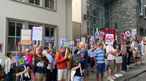 The image shows protesters standing outside Guernsey's Royal Court. There is an assortment of men and women holding placards with anti-GST signs on them. They are all standing on the pavement next to a road. 