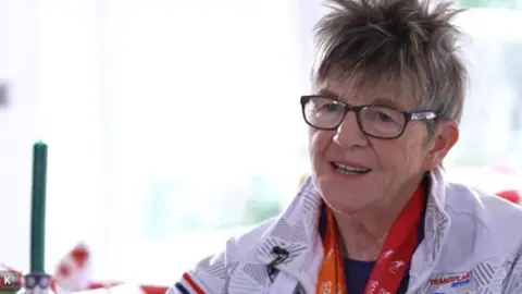 NHS Blood and Transplant handout Nicki Detko with grey and blond short hair wearing black glasses and a Team GB white tracksuit and medals from the Transplant Games. She is sitting in front of a window.