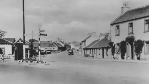 A black and white image of a small Scottish village with houses and a road sign in the middle