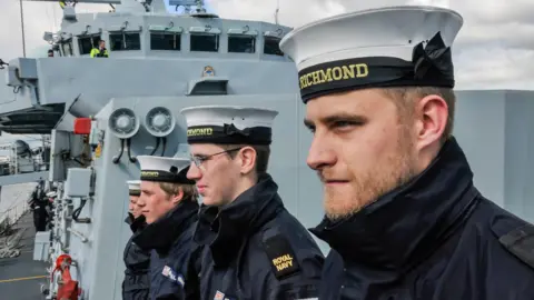 Four sailors, in white hats which read Richmond on the front, stand in a line on board a Navy ship. 