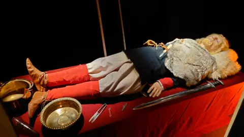 Discover Bucks Museum A replica of a Saxon burial showing a mannequin of a man's body with long blond hair ahead of its burial. It dressed Saxon clothing with replicas of its grave goods including a sword, two bowls, tweezers, shears and a drinking horn.