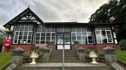A black and white half-timber building at the top of two flights of steps. It is set within a park, with a lawned embankment in front and trees behind.