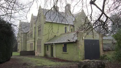 A side view of a large, boarded-up Victorian building. The weather is grey and overcast.