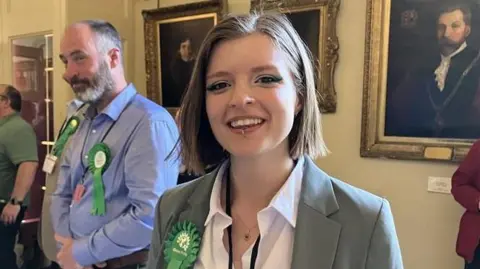 A woman with neck-length brown hair and a lip piercing smiles at the camera. She is wearing a white blouse under a grey jacket with a green rosette pinned to her lapel.