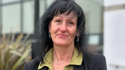 Jackie Hallam, who has long dark hair and is wearing a black jacket over a green top.