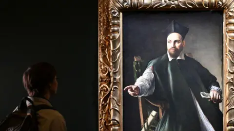 Getty Images Young person with backpack looks at framed portrait of bearded cleric with a black tricorn hat wearing a dark robe, his right hand outstretched and his left hand clutching a piece of paper