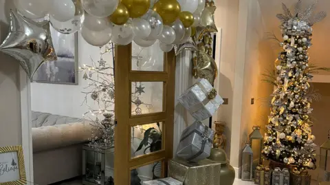 IWC Media A Christmas themed hallway. Gold and white balloons and a stack of gold and white presents border a living room door. In the corner is a tall Christmas tree decorated with white and gold Christmas baubles and lit up in warm white fairy lights. 