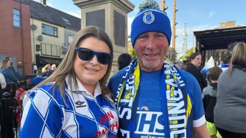Katherine McCausland and Roy Stretton at the Dungannon Swifts homecoming. Katherine has long brown hair, wearing sunglasses and blue and white jersey. Roy is wearing a blue bobble hat, blue t-shirt and Dungannon scarf.