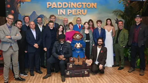 A group photo of a number of Paddington in Peru cast and crew members, including Olivia Colman, Ben Whishaw, Hugh Bonneville, and Emily Mortimer. In the centre of the group shot, there is a replica of Paddington Bear, who is sat on top of three stacked suitcases, eating a marmalade sandwich.