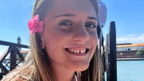 Juliette, smiling at the camera on a sunny day, is wearing her long hair down, sunglasses on her head and a pink flower tucked above her ear. She is sitting by the water